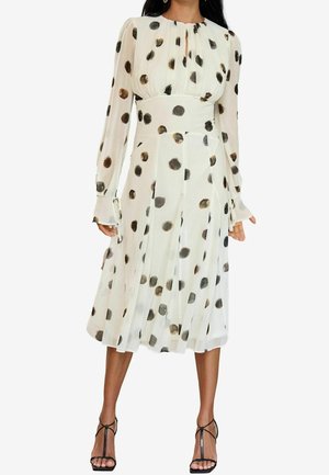 Kobieta ubrana w białą sukienkę midi z długimi bufkami i rozmieszczonymi dużymi czarnymi oraz brązowymi groszkami, w połączeniu z czarnymi sandałami na obcasie z paskami.