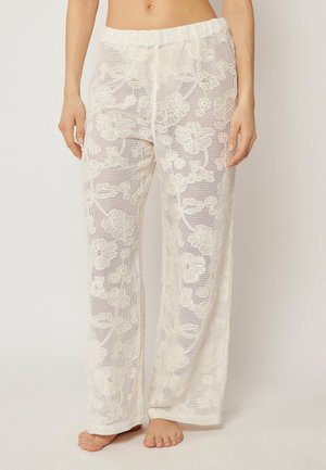 Pantalon large blanc transparent avec motif en dentelle florale, taille élastique et pieds nus visibles de la taille jusqu'aux pieds.