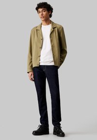 Chaqueta abotonada verde oliva, camiseta de cuello redondo blanca, jeans de mezclilla azul oscuro y zapatos negros de cordones; tejido suave, corte informal y diseño sencillo.