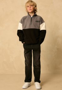 Grå og sort fleece-pullover med hvide detaljer, quarter-zip, parret med mørke jeans og hvide sneakers, stående mod en neutral baggrund.