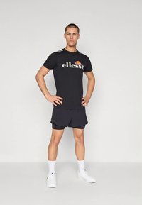 Svart atletisk t-skjorte med «ellesse»-logo, kombinert med svarte shorts og hvite joggesko. Enkel, minimalistisk design med korte ermer.