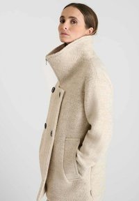 Abrigo de lana beige con cuello alto, botones de doble botonadura, bolsillos laterales y una silueta estructurada, que presenta una tela suave y texturada.