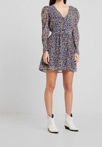 Jurk met bloemenprint op een marineblauwe achtergrond, met poflange mouwen, V-hals, getailleerde taille en uitlopende rok. Draagt witte enkellaarsjes.