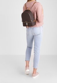 Hnědý batoh s monogramem a béžovými akcenty, se zipem a nastavitelnými popruhy. Na sobě má růžový svetr a světlé džíny s casual obuví.