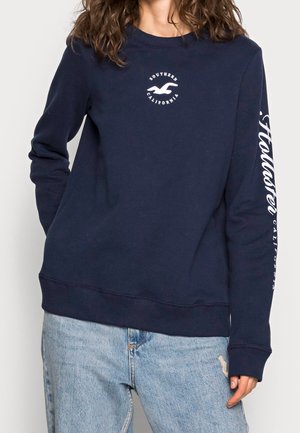 Personne portant un sweat-shirt bleu marine avec le texte et le logo "Southern California" sur la poitrine et "Hollister California" imprimé sur la manche droite.