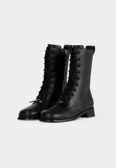 Paire de bottes montantes en cuir noir à lacets, fermetures éclair latérales et petits talons blocs sur un fond clair uni.