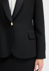 Černý blazer vyrobený z hladké tkaniny, s saténovými klopami, zlatým zapínáním na knoflík a malou kapsou s lemovaným okrajem.