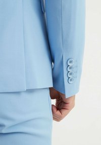 Costume bleu clair avec une finition texturée ; le poignet de la veste présente quatre boutons ronds de la même couleur que le tissu, mettant en valeur un design sur mesure.