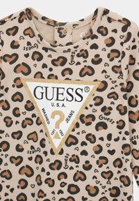 Beige lange mouwen shirt met luipaardprint, voorzien van zwarte en bruine vlekken. Heeft een driehoekige, goudaccentuerde "GUESS" logo in het midden.