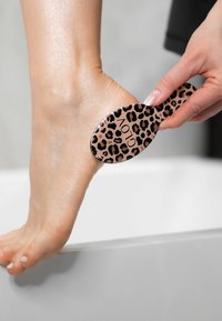 Spazzola per piedi con stampa leopardata, dotata di una testa ovale liscia e un manico corto, utilizzata per la cura dei piedi, tenuta contro il piede di una persona vicino a una superficie bianca.