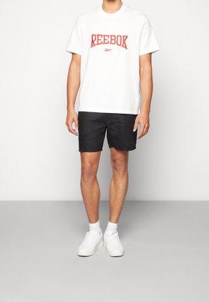T-shirt en coton blanc avec le logo "Reebok" rouge, assortie à un short noir et des baskets blanches. Le modèle se tient sur un fond gris neutre.