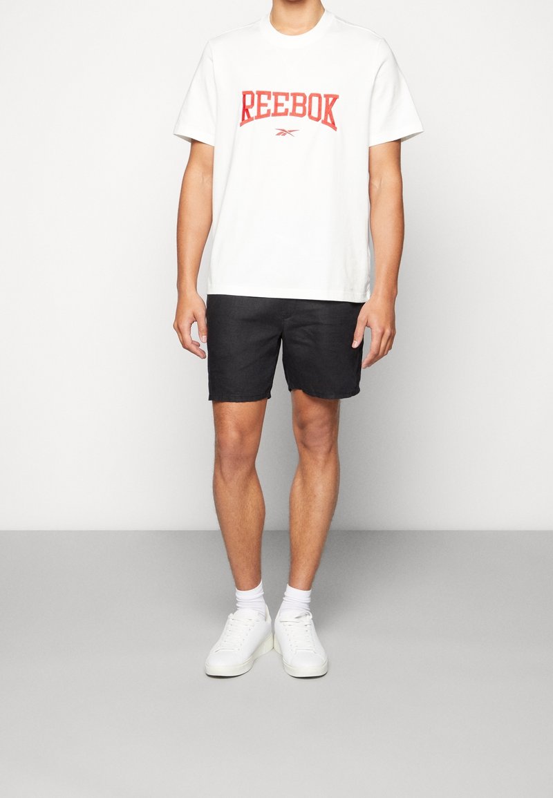 Vitt bomullströja med röd "Reebok"-logga, kombinerat med svarta shorts och vita sneakers. Modellen står framför en neutral grå bakgrund.