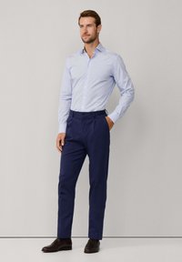 Uomo che indossa una camicia da uomini azzurro chiaro, pantaloni su misura blu navy e mocassini marroni scuri, in piedi contro uno sfondo chiaro e semplice.