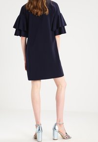 Marineblaues Kleid mit ausgebelltem Ärmel, knielang. Silberne High-Heels mit Knöchelriemen. Glatte Stofftextur, minimalistische Details.