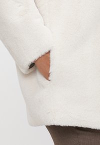 White faux fur jacket with a soft texture, featuring a visible pocket and a slightly oversized design. Hemline is straight with no additional detailing.