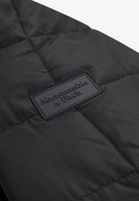 Must musta jakk, millel on hõbedane brändilogo silt, klapid ja sile, läikiv kangas. 