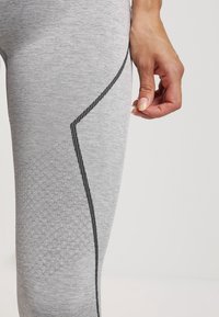 Szare legginsy sportowe mają teksturowany wzór na dolnej części nogawki oraz kontrastującą czarną szew wzdłuż boku, zapewniając dopasowany kształt.