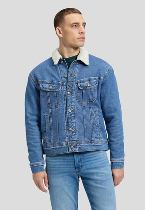 Homme debout, portant une veste en jean bleu avec un col sherpa blanc et un jean bleu assorti, légèrement tourné vers la gauche devant un fond uni.