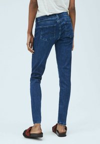 Žena v tmavě modrých skinny džínách a bílém tričku, stojící s jednou rukou uvolněně podél těla, obutá v pletených sandálech na slip.