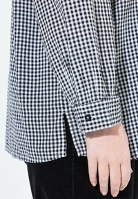 Schwarz-weißer karierter Hemd mit geknöpften Manschetten und seitlichen Schlitzen, aus Baumwollstoff. Nahaufnahme von Ärmel und Handdetail.
