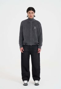 Junger Mann mit dunklen Haaren, der einen grauen Reißverschluss-Strickpullover, lockere schwarze Hosen und schwarz-weiße Sneaker trägt, steht vor einem weißen Hintergrund.