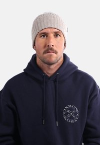 Un bonnet en tricot côtelé beige porté avec un sweat à capuche bleu marine présentant un imprimé logo circulaire. Le bonnet a un ajustement serré et une texture intéressante.