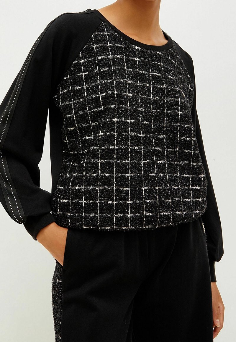 Haut noir à manches longues avec un panneau avant texturé présentant un motif de grille blanche, associé à un pantalon noir, mains dans les poches.