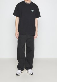 Camiseta de algodón negra con mangas cortas y un gráfico verde en el lado izquierdo del pecho, combinada con pantalones negros de corte holgado y zapatillas grises.