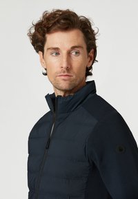 Veste marine avec un devant matelassé gonflé, un tissu lisse sur les épaules, col haut et un logo en accent sur le bras gauche, dotée d'un zip.
