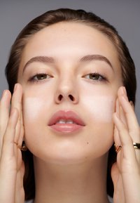 Ansikt med påført hvit krem; glatt tekstur, lys hudtone, plasserte hender, minimal sminke, naturlig lys. Nøytral bakgrunn.