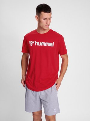 Mann trägt rotes hummel T-Shirt und hellgraue hummel Shorts, steht mit den Händen in den Taschen und schaut nach links vor einem einfarbigen Hintergrund.