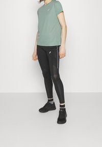 Vihreä lyhythihainen urheilu paita, joka on yhdistetty mustiin leggings-hoosiin, joissa on verkkopaneeleja ja valkoisia yksityiskohtia, käytetty mustien urheilukenkien kanssa.
