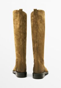 Bottines en daim marron, arrivant au genou, avec une texture lisse, semelle en caoutchouc noir plat et coutures minimales à l'arrière et sur les côtés.