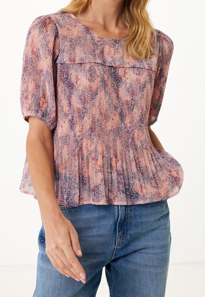 Femme portant un chemisier plissé à manches courtes avec des motifs abstraits violets et pêche, associé à un jean bleu, fond blanc.