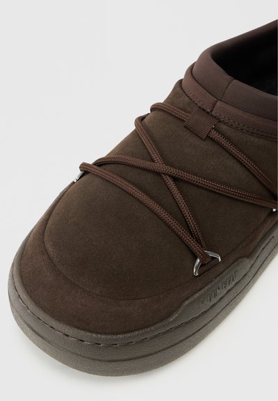 Zapato de ante marrón tipo mocasín con punta redonda, cordones elásticos, suela de goma y detalles de costura sutiles. Textura suave con un acabado mate.