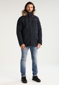Navy modrá parka s kapucí lemovanou kožešinou, obsahující zipové kapsy, červené akcenty a volný střih. Materiál je odolný vůči počasí.