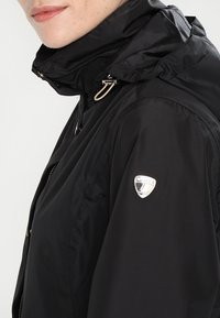 Giacca nera leggera con collo alto, chiusura con cerniera e dettaglio in metallo argentato sulla manica. Texture liscia e design minimalista.