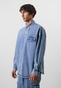 Camisa de ganga oversized azul clara com fecho de botões, bolso no peito e mangas longas com punhos. Bainhas desgastadas e textura macia.