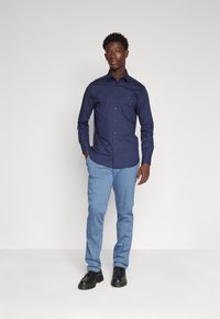 Camicia a maniche lunghe blu navy con chiusura a bottoni, abbinata a pantaloni blu chiaro e scarpe nere, con un logo discreto sulla camicia.