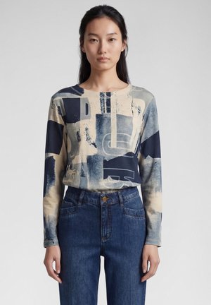 Frau mit langen dunklen Haaren, die ein langärmliges, abstrakt gemustertes Hemd und blaue Jeans trägt, steht vor einem schlichten hellen Hintergrund.