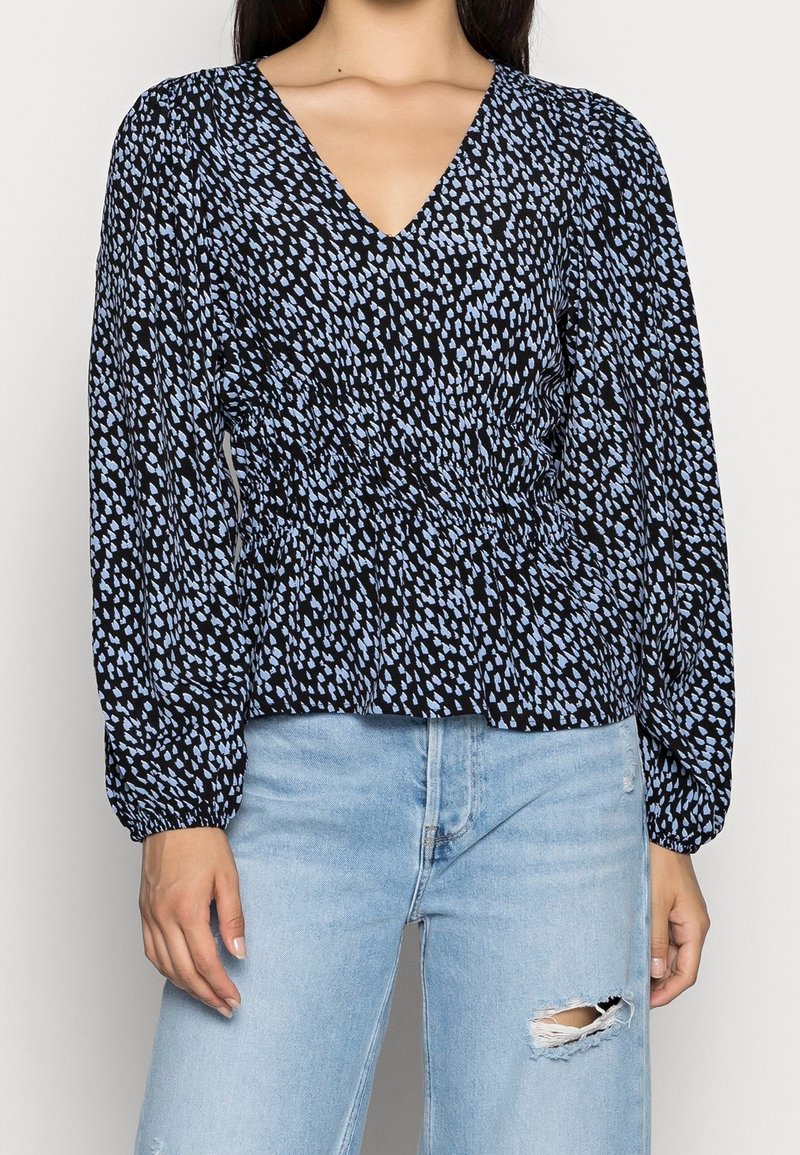 Zwarte blouse met blauwe abstracte print, V-hals, lange mouwen en een geplooide taille, gecombineerd met lichtblauwe jeans met slijtageplekken.