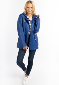 Blaue wasserdichte Jacke mit Kapuze, goldener Hardware, Frontreißverschluss und Druckknöpfen. Kombiniert mit dunklen Jeans und weißen Sneakern.