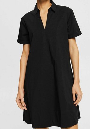 Femme portant une robe noire ample à manches courtes avec un col en V, debout devant un fond clair uni.