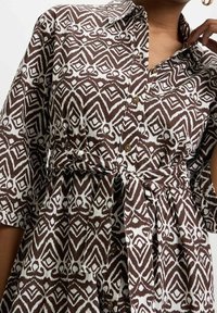 Brun och vit mönstrad skjortklänning med krage, knäppning fram, korta ärmar och knytbar midja, med ett geometriskt mönster.