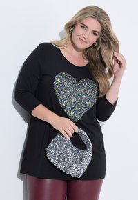 Tunique noire avec un grand design de cœur en sequins multicolores, accompagnée d'un sac à main en sequins argentés brillant et de leggings bordeaux.
