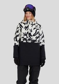 Donna che indossa una giacca da sci a righe bianche e nere, pantaloni da neve neri, guanti neri, un berretto nero e occhiali da sci viola sulla testa.