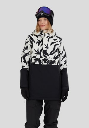 Frau trägt eine schwarz-weiße gemusterte Ski-Jacke, schwarze Schneehose, schwarze Handschuhe, eine schwarze Mütze und lila Skibrille auf dem Kopf.