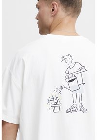 Hvid bomulds t-shirt med en sort stregtegning af en person, der vander en blomst i potte på ryggen. Korte ærmer, afslappet pasform.