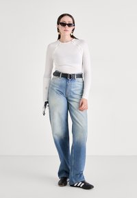 Weißes Langarmoberteil mit dekorativen Knöpfen, hellblaue weit geschnittene Jeans, schwarzer Gürtel und schwarze Sonnenbrille. Schwarz-weiße Slipper.