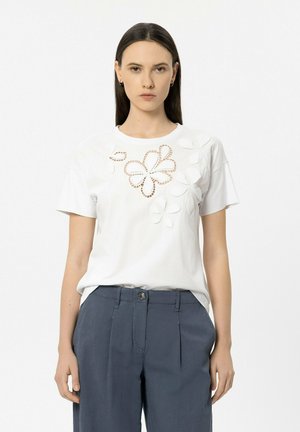 Frau trägt ein weißes Kurzarmhemd mit floralen Cut-outs und Applikationsdetails, kombiniert mit hoch taillierten, dunkelblauen, plissierten Hosen.